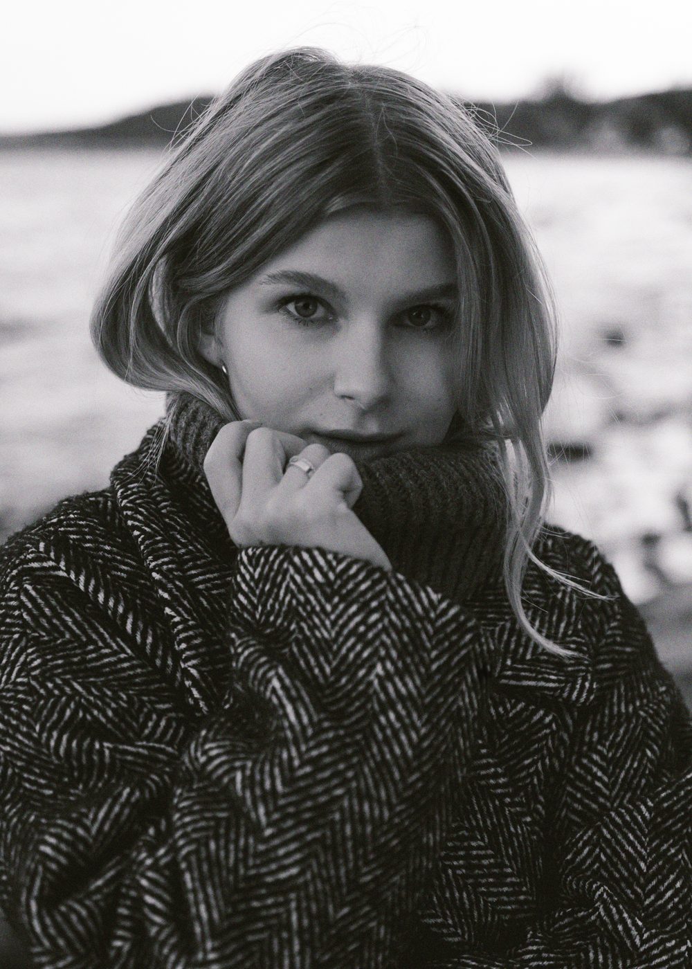 Close portrait of a woman pulling a herringbone coat collar up to her chin by the water