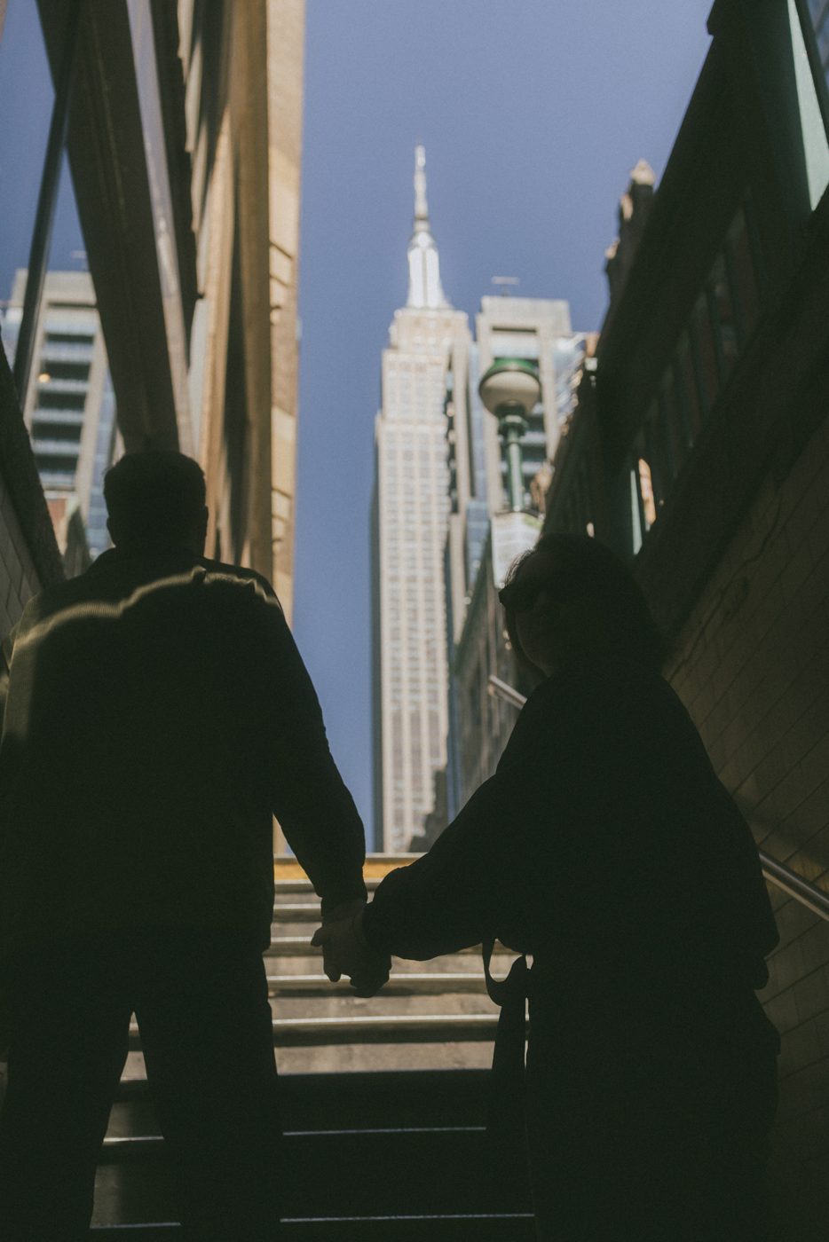 Couple in silhouette holding hands on subway stairs, the Empire State Building above them