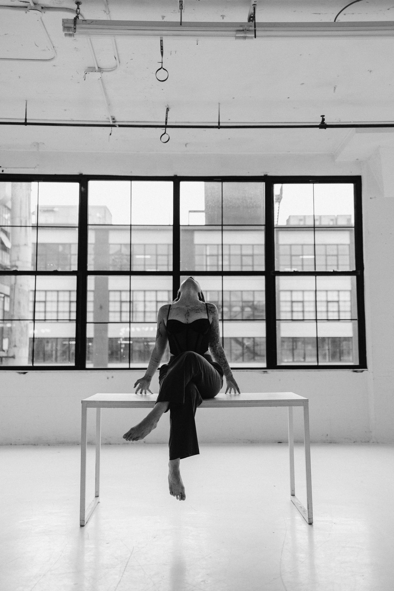 Woman sitting on a white table in an empty loft, head tilted back, large factory windows behind her