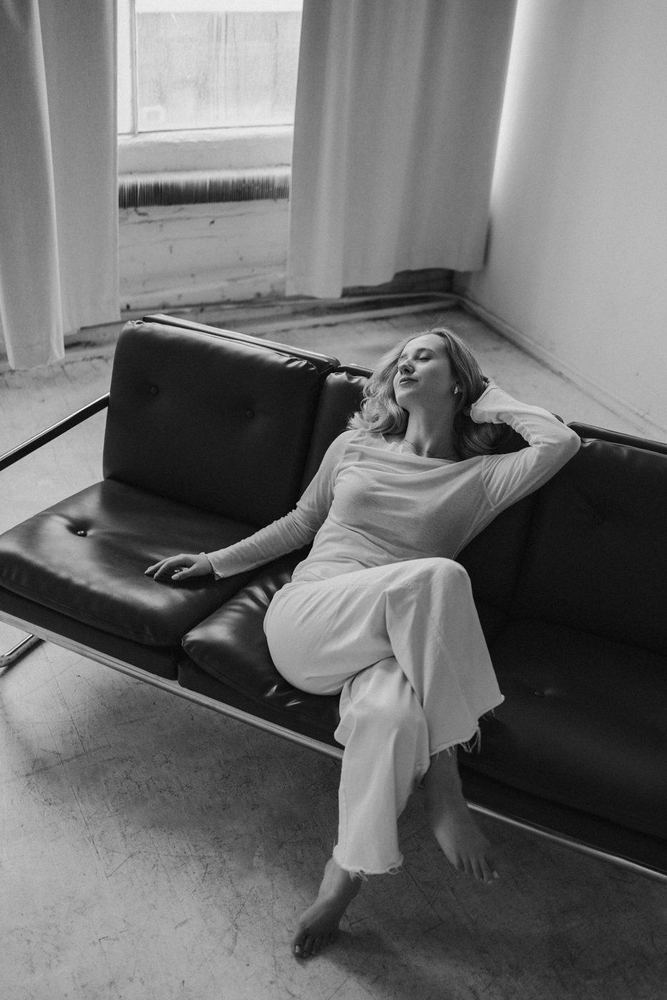 Woman in white reclining on a black leather sofa, eyes closed, soft window light