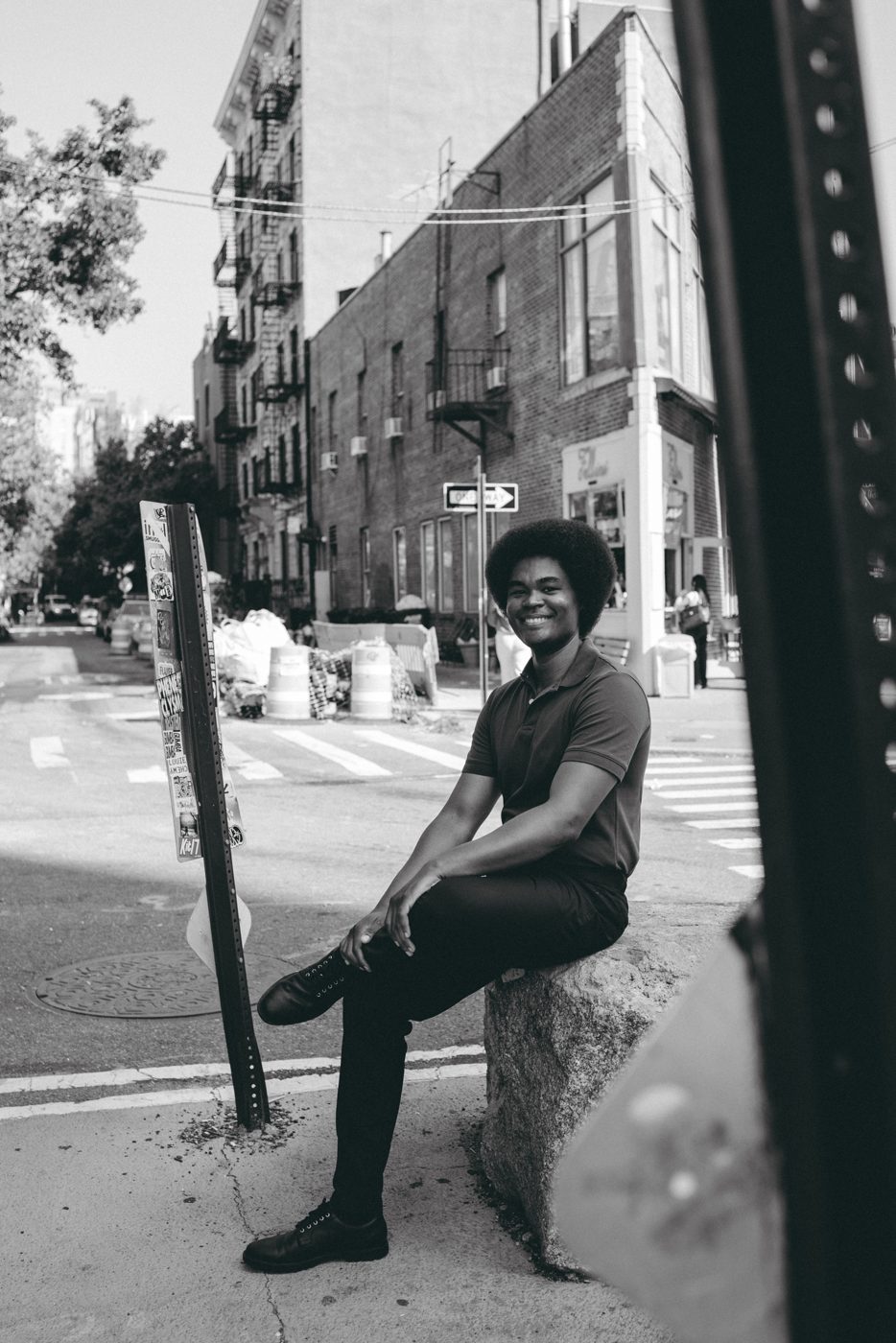 Smiling man sitting on a rock at a New York street corner, framed by a signpost